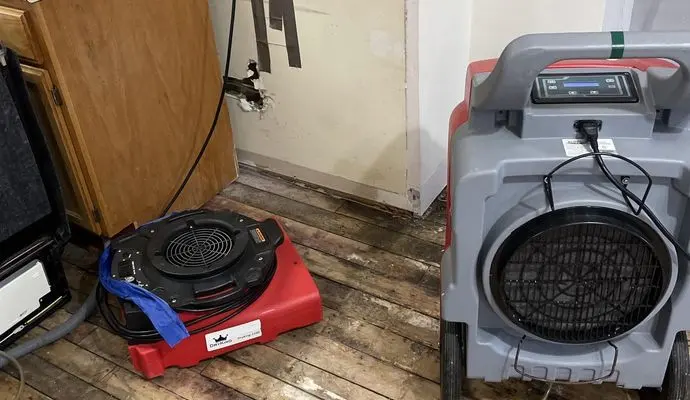 Oregon flood season water damage: an air mover and a dehumidifier running on a stripped wood floor beside kitchen cabinets